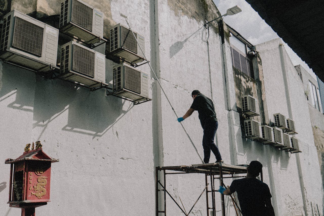 Two workers on scaffold maintain air conditioning units attached to a building, displaying urban upkeep.